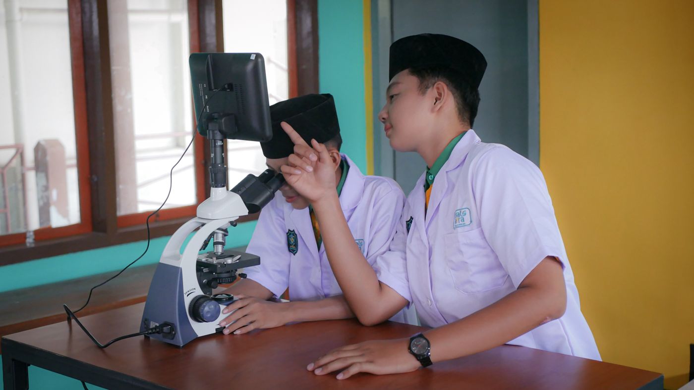 Dua siswa berjas lab mengamati preparat dengan mikroskop digital